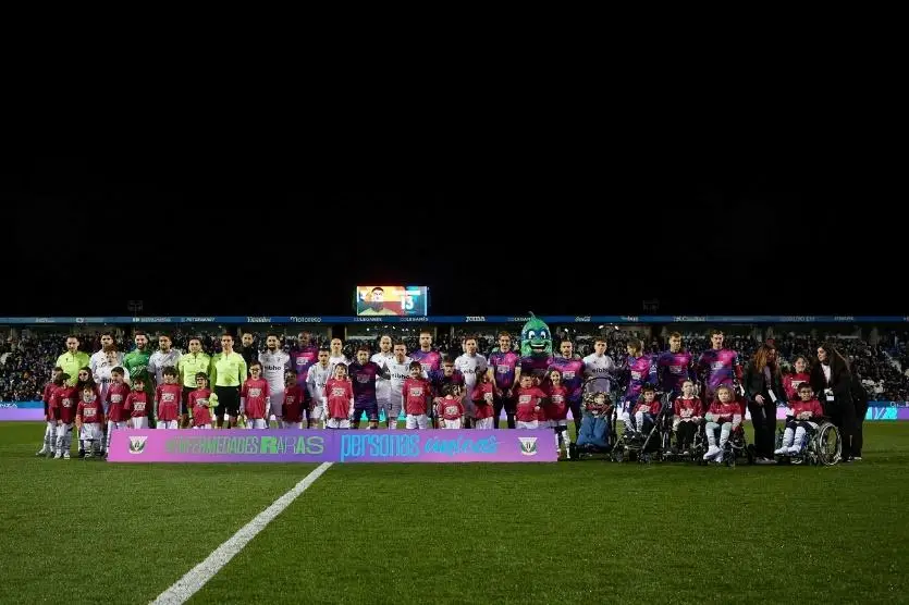 I giocatori di due squadre di calcio posano con dei bambini, alcuni in sedia a rotelle, dietro uno striscione di sensibilizzazione sulle malattie rare.