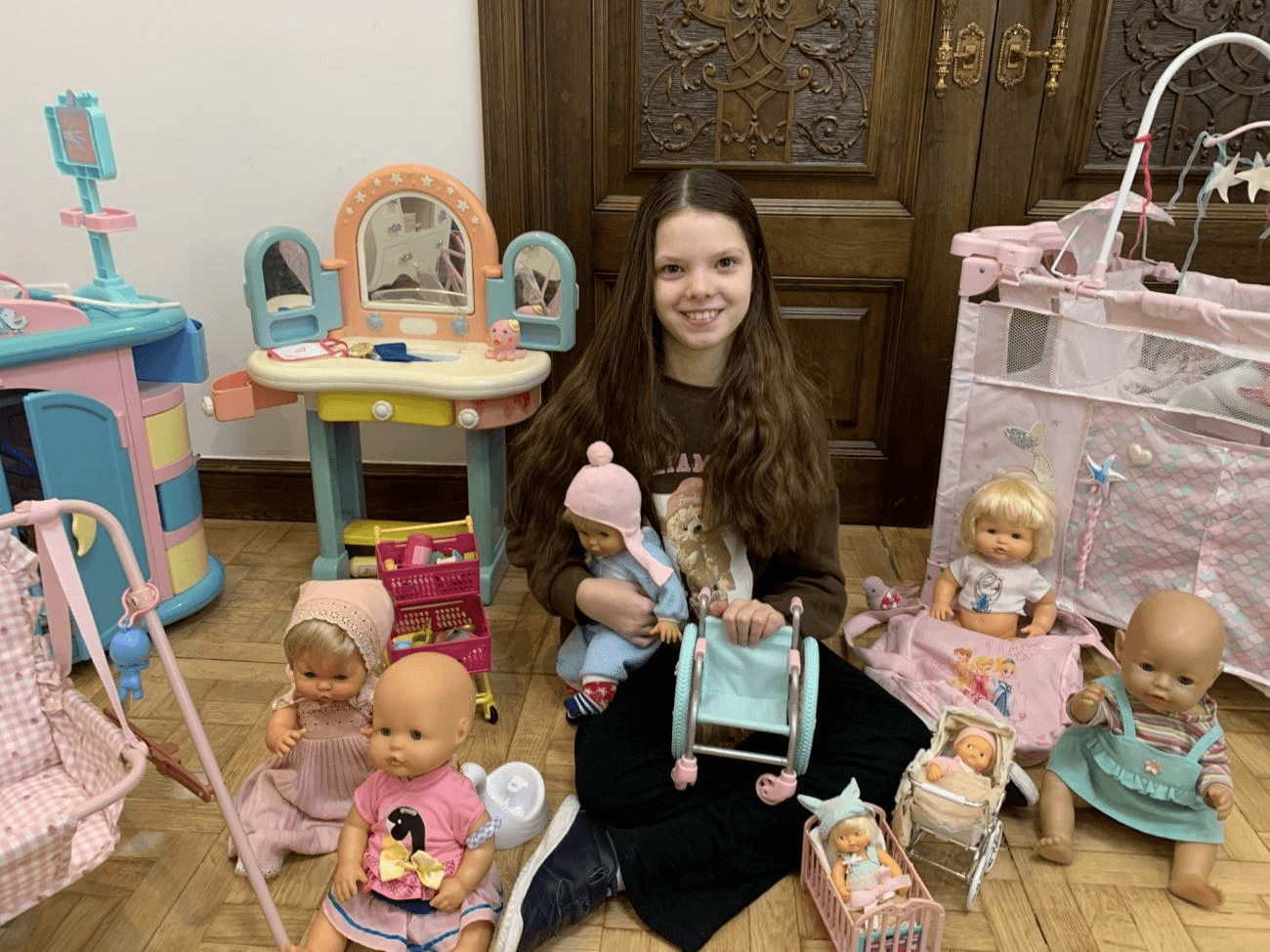 Una bambina sorride seduta per terra, circondata da bambole e giocattoli, tenendo in mano una sedia a rotelle giocattolo.