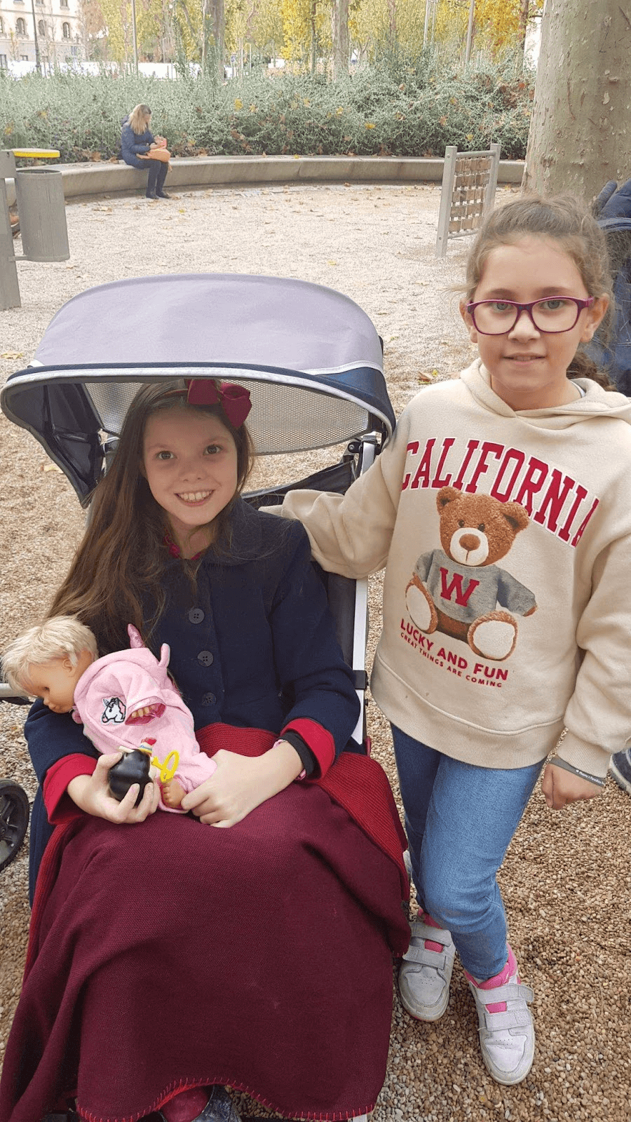 Carolina, sulla sua sedia adattata e con una bambola in braccio, nel parco accanto alla sua migliore amica Patricia.