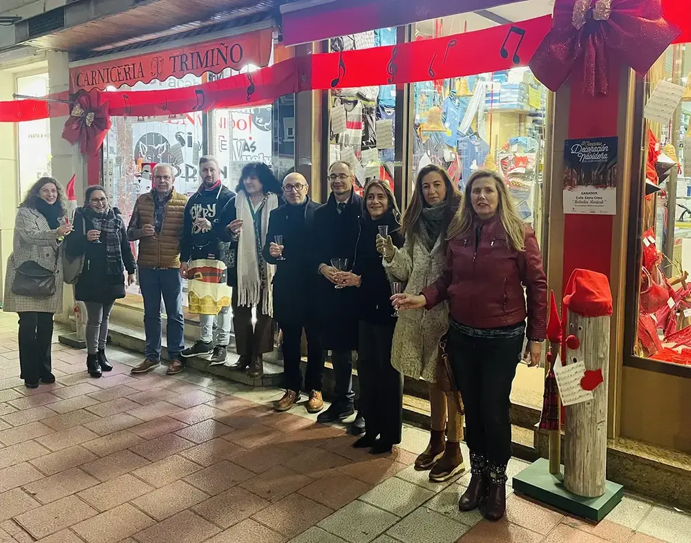 Un gruppo brinda con i calici davanti a un negozio decorato con fiocchi rossi e note musicali, accanto alla locandina del concorso natalizio.