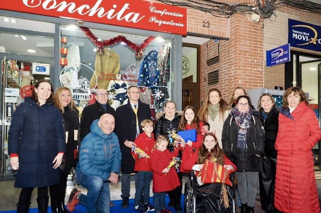 Foto di gruppo davanti al negozio Conchita, con diversi bambini con il cappotto rosso e una bambina in sedia a rotelle in primo piano.