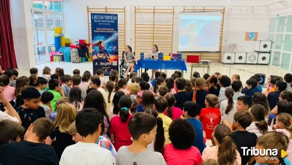 Decine di scolari seduti per terra ascoltano due bambine, una in sedia a rotelle, accanto al roll-up del libro.