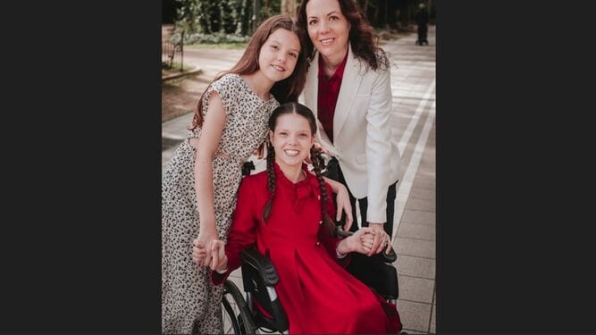 Ritratto all'aperto: Carolina, vestita di rosso sulla sua sedia a rotelle, tiene per mano la sorella e la madre, che la abbracciano.