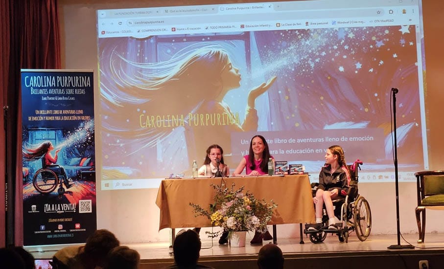 Presentazione in un'aula magna: la copertina proiettata, il roll-up del libro e una bambina in sedia a rotelle accanto al tavolo.