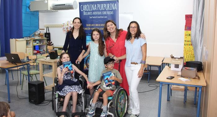 Foto di gruppo in un'aula davanti al roll-up del libro: tre donne, una bambina e due minori in sedia a rotelle con delle copie.