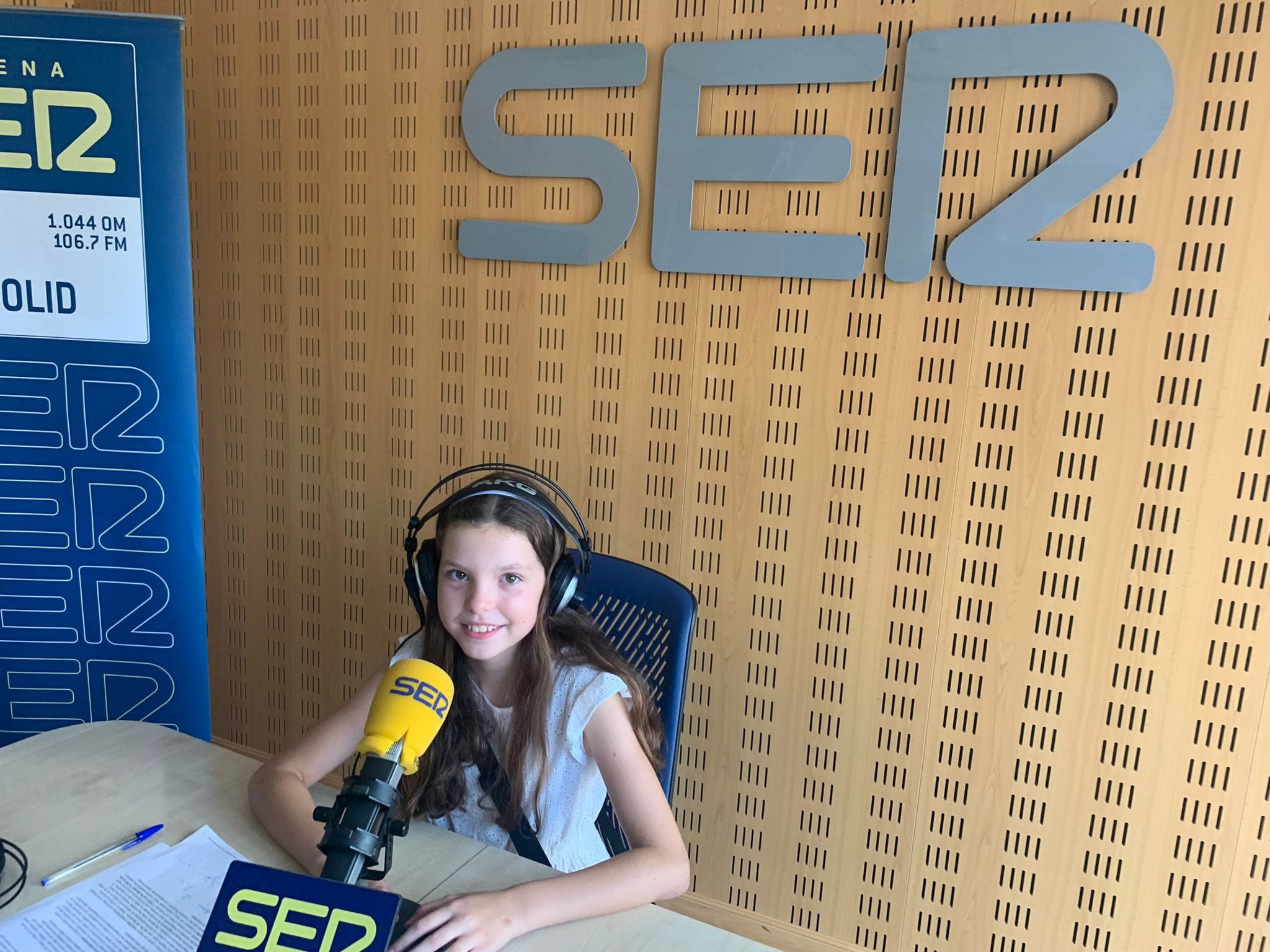 Una bambina con le cuffie sorride davanti al microfono giallo della Cadena SER, sotto il logo dello studio.
