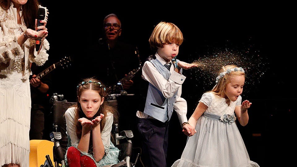 Sul palco, tre bambini soffiano glitter; una di loro, in sedia a rotelle, porta una corona di fiori.