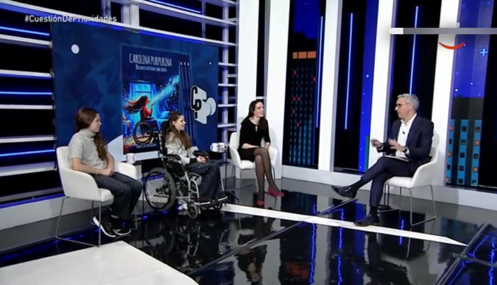 Studio televisivo: due ragazze, una in sedia a rotelle, e una donna conversano con il presentatore davanti alla copertina del libro.