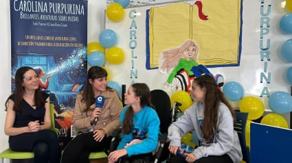 Intervista di COPE in una scuola: una giornalista con il microfono accanto a Carolina e ad altre due ragazze, davanti al roll-up del libro.