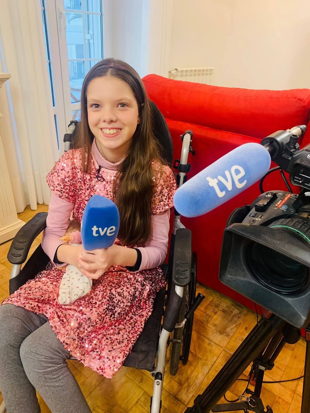 Carolina, sulla sua sedia a rotelle e con un vestito rosa di paillettes, tiene un microfono di TVE davanti alla telecamera.