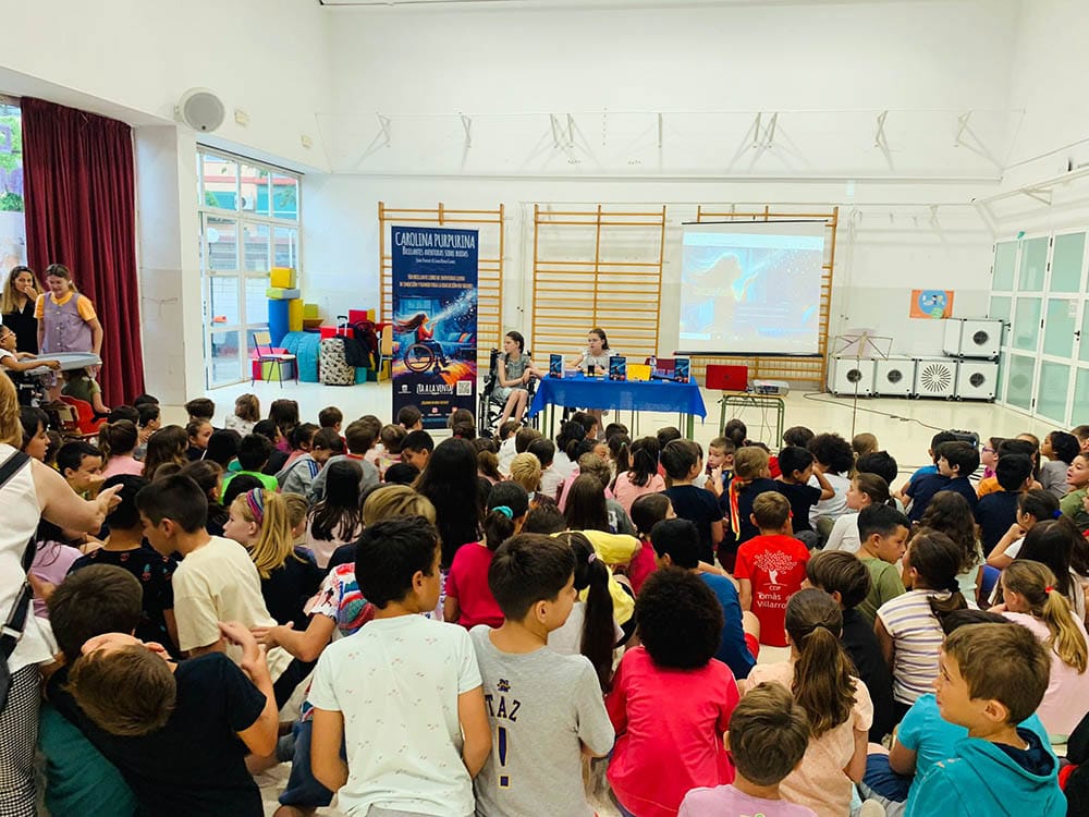 Decine di scolari seduti per terra ascoltano due bambine al tavolo, una in sedia a rotelle, accanto al roll-up del libro.
