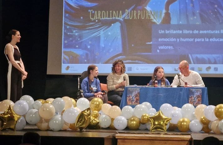 Dibattito sul palco di una scuola: quattro persone al tavolo, una al microfono, tra palloncini dorati e bianchi.