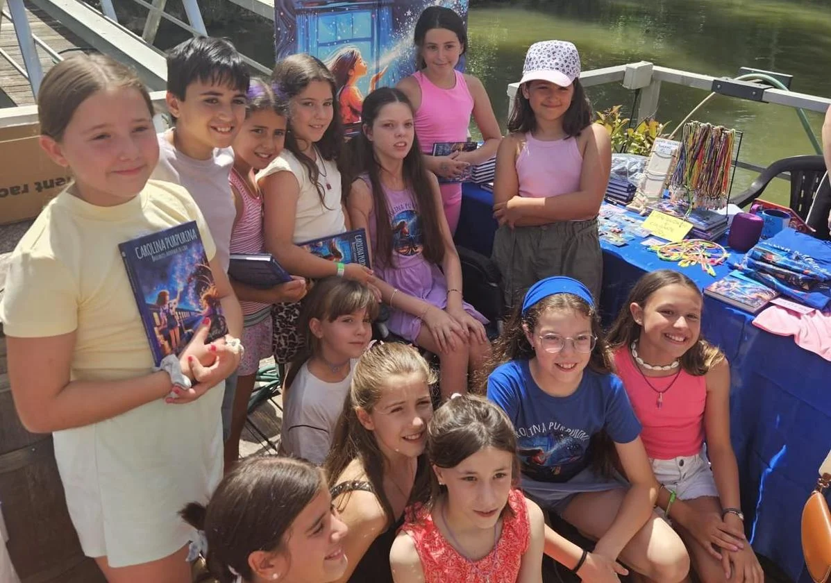 Un gruppo di bambine posa accanto al banchetto di vendita in riva al fiume; alcune tengono il libro «Carolina Purpurina».