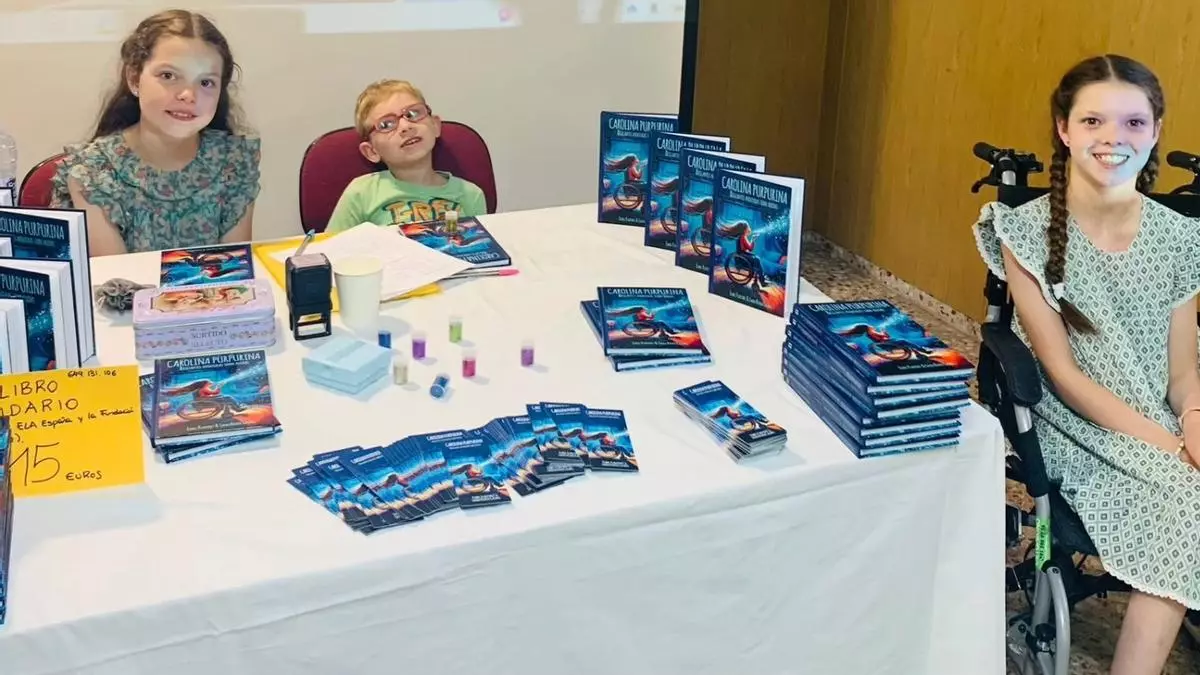 Tavolo delle firme con pile del libro, segnalibri e tubetti di glitter; a destra, una bambina in sedia a rotelle.