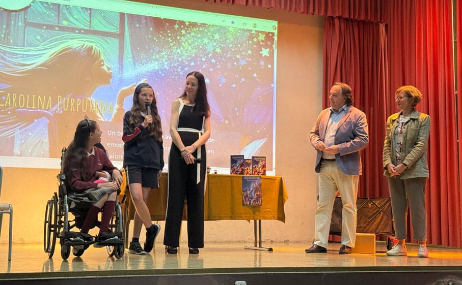 Sul palco, un'adolescente parla al microfono accanto a Carolina sulla sua sedia a rotelle, con la copertina proiettata sullo sfondo.
