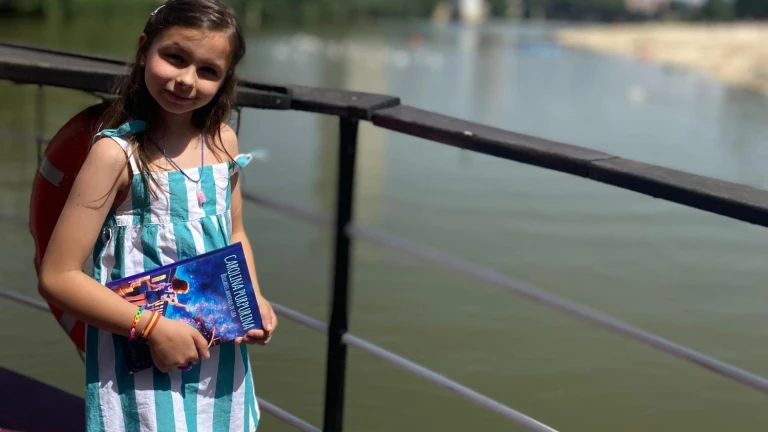 Una bambina con un vestito a righe e uno zaino tiene una copia di «Carolina Purpurina» accanto alla ringhiera di un fiume.