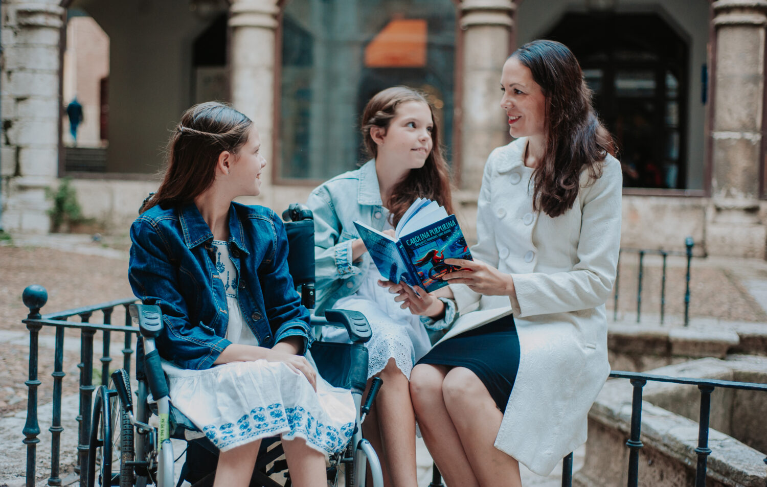 Laura legge il libro a Carolina, sulla sua sedia a rotelle, e a Isabel in un cortile antico.