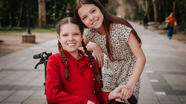 Carolina, sulla sua sedia a rotelle, sorride mentre un'altra bambina si appoggia alla sua spalla in un viale alberato.