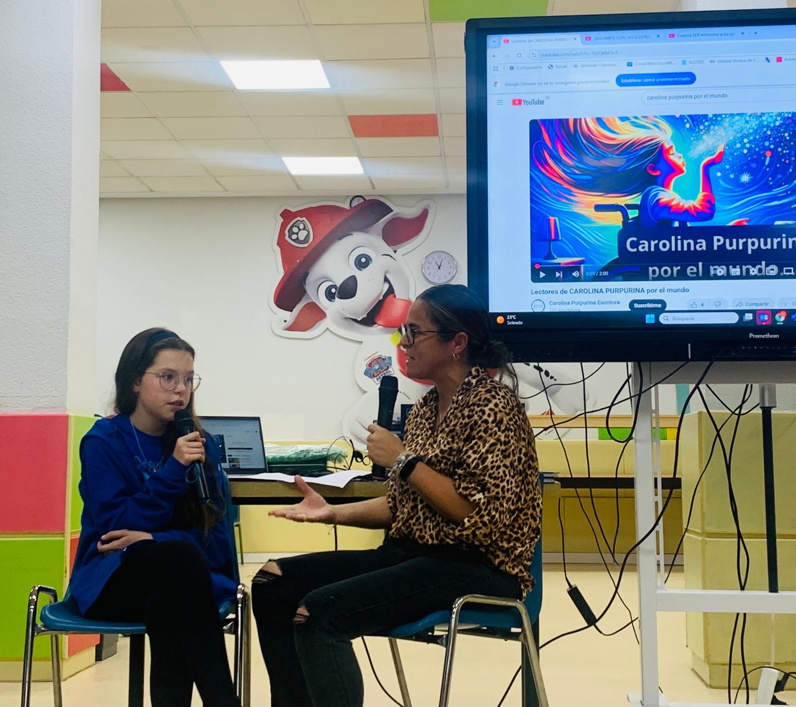 Isabel, con il microfono, risponde a un'intervistatrice davanti a uno schermo che proietta il video «Carolina Purpurina por el mundo».