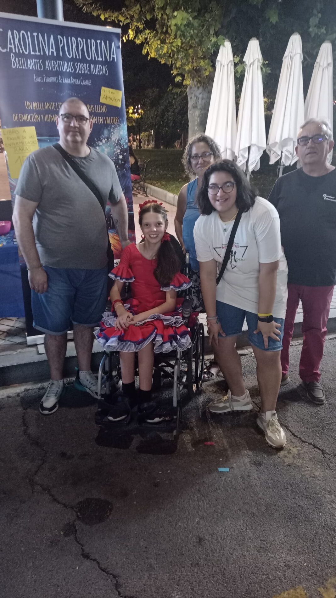 Di sera alla fiera, una bambina in abito da flamenca e in sedia a rotelle posa con diverse persone davanti al roll-up del libro.