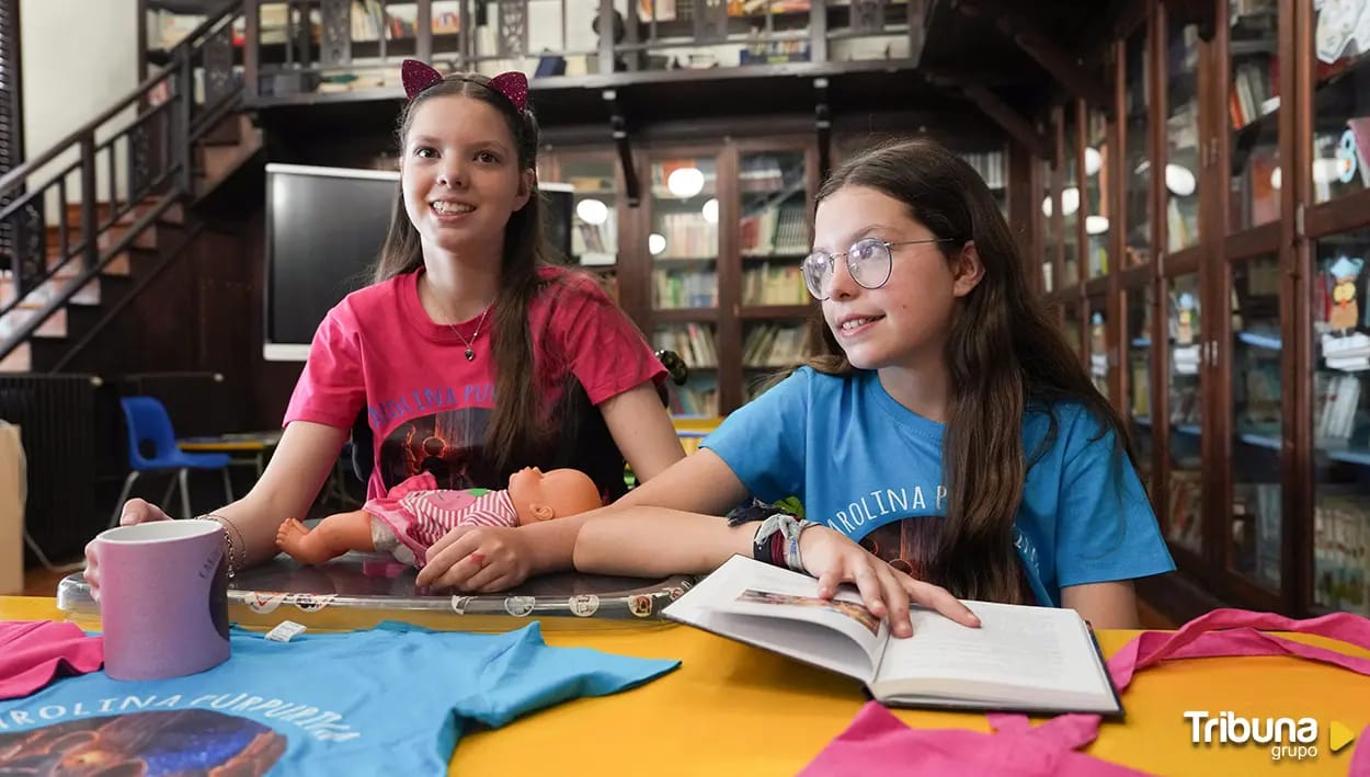 Carolina e Isabel in una biblioteca, con il libro aperto, una bambola e magliette di Carolina Purpurina.