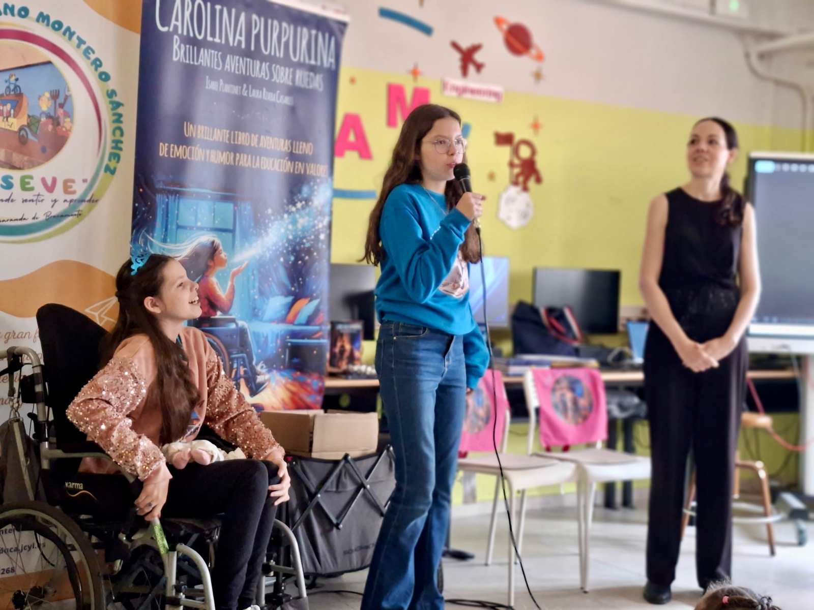 Isabel parla al microfono durante l'incontro scolastico, con Carolina e Laura al suo fianco.