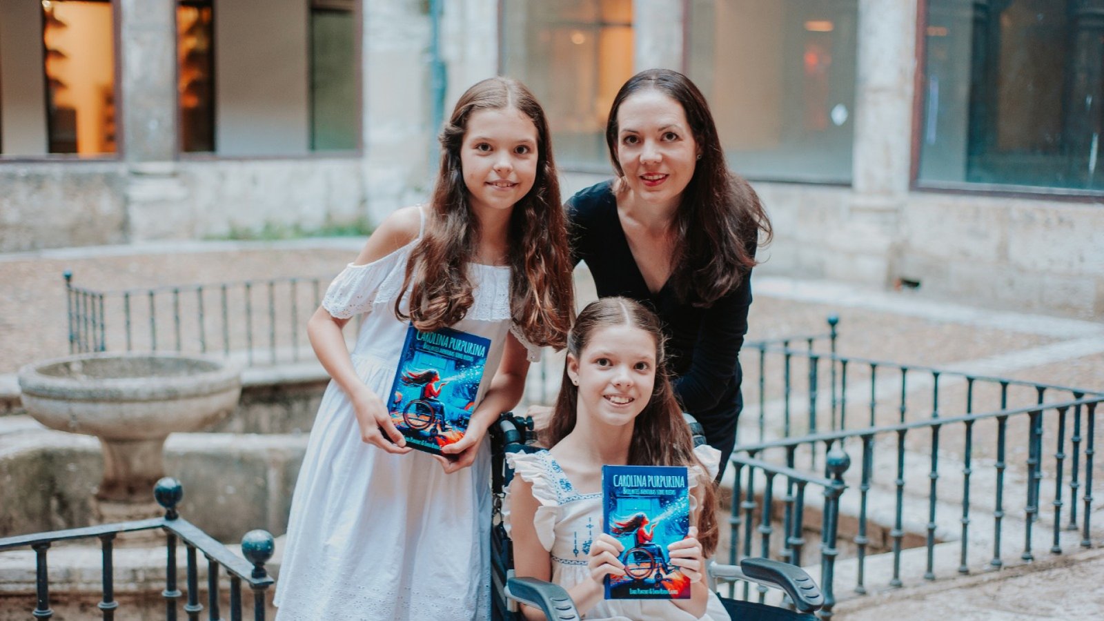 Carolina, sulla sedia a rotelle, e un’altra bambina mostrano il libro «Carolina Purpurina» accanto a una donna, in un cortile di pietra.