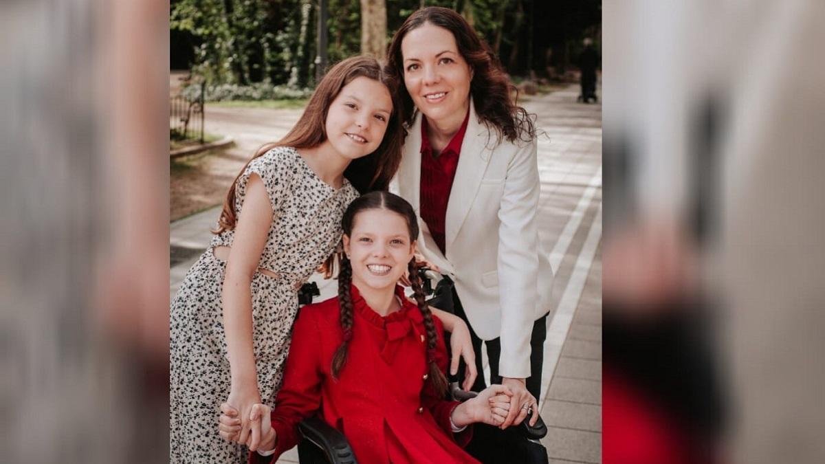 Carolina, con abito rosso e trecce, sorride sulla sua sedia a rotelle, tra una bambina con abito a fantasia e una donna in giacca bianca.