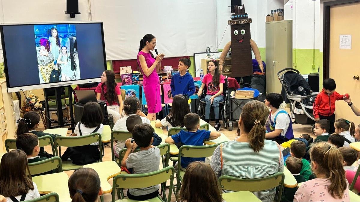 Una donna parla al microfono davanti agli alunni seduti; accanto a lei alcuni bambini e Carolina, sulla sedia a rotelle, vicino a un tavolo di libri.