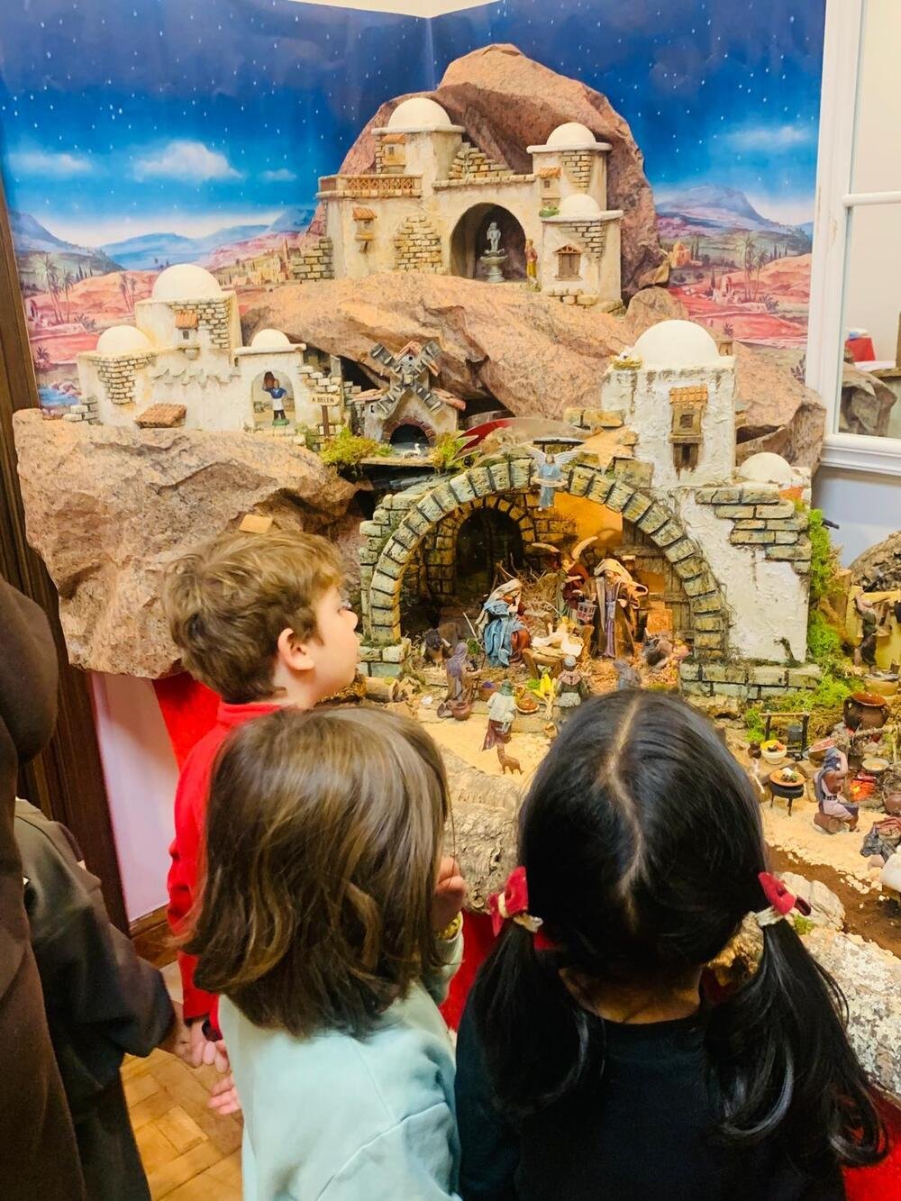 Tre bambini, di spalle, guardano il presepe: case bianche, una grotta in pietra con la Natività e un fondale di cielo stellato.