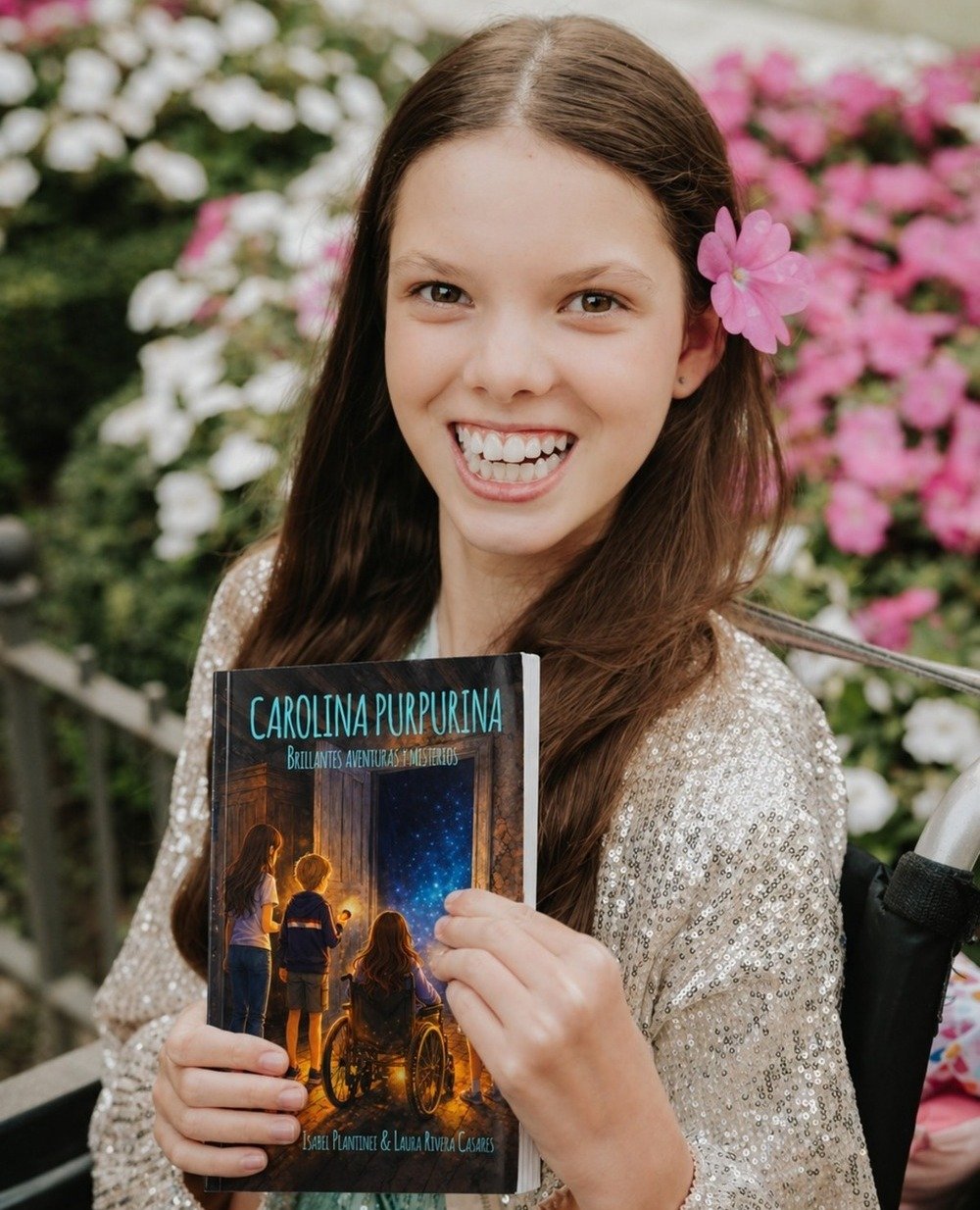 Una bambina sorridente, con un fiore rosa tra i capelli, tiene il libro «Carolina Purpurina. Brillantes aventuras y misterios».