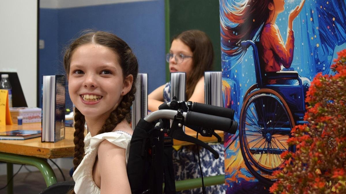 Presentazione del libro: Carolina sorride sulla sua sedia a rotelle accanto al roll-up della copertina; dietro, una bambina seduta al tavolo dei libri.