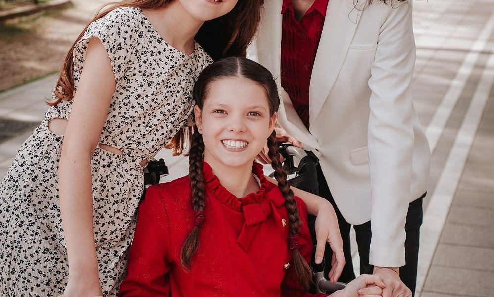 Carolina, con abito rosso e trecce, sorride sulla sua sedia a rotelle, abbracciata dalla famiglia, i cui volti restano fuori dall’inquadratura.