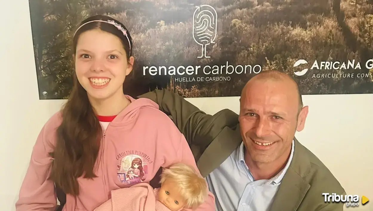 Una bambina con la felpa rosa di Carolina Purpurina e una bambola in braccio posa con un uomo davanti al pannello di Renacer Carbono.